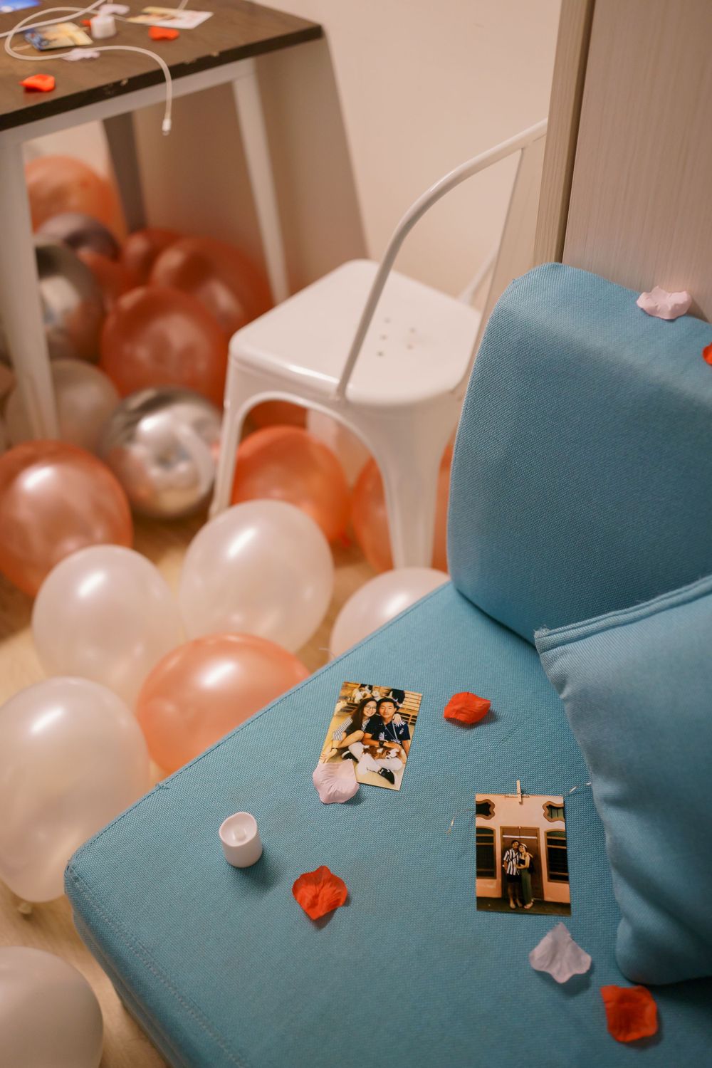 Party decorations including rose gold and white balloons scattered near a blue couch with small photos and petals.