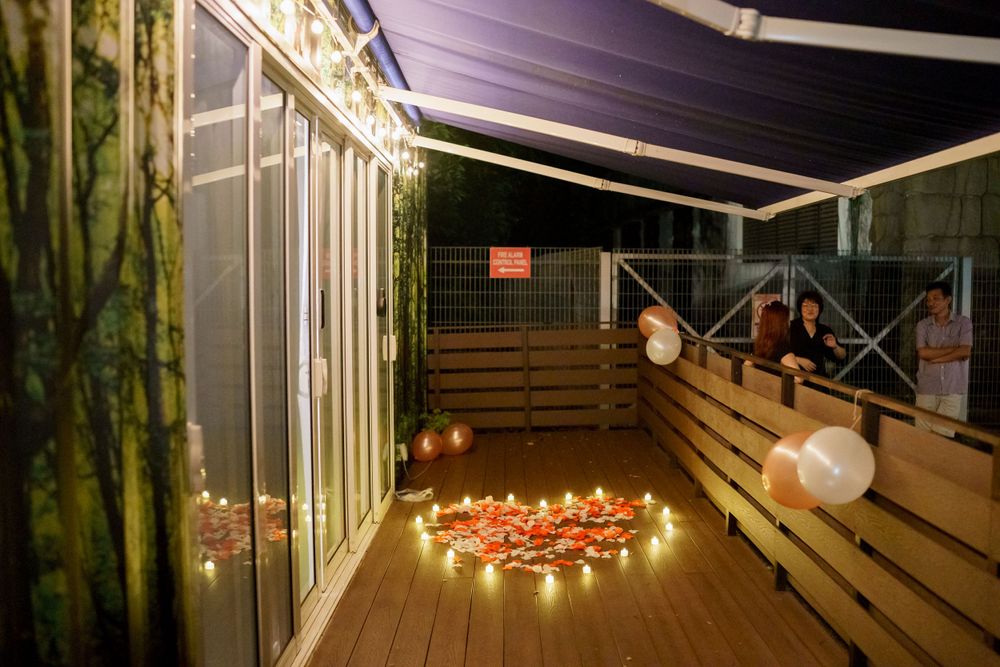 Tea lights arranged in a heart shape on a wooden deck at night with paper lanterns and string lights creating ambiance.
