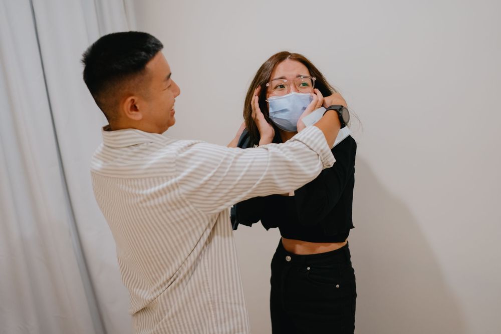 Healthcare worker adjusts protective face mask on patient during medical examination against light curtain background.