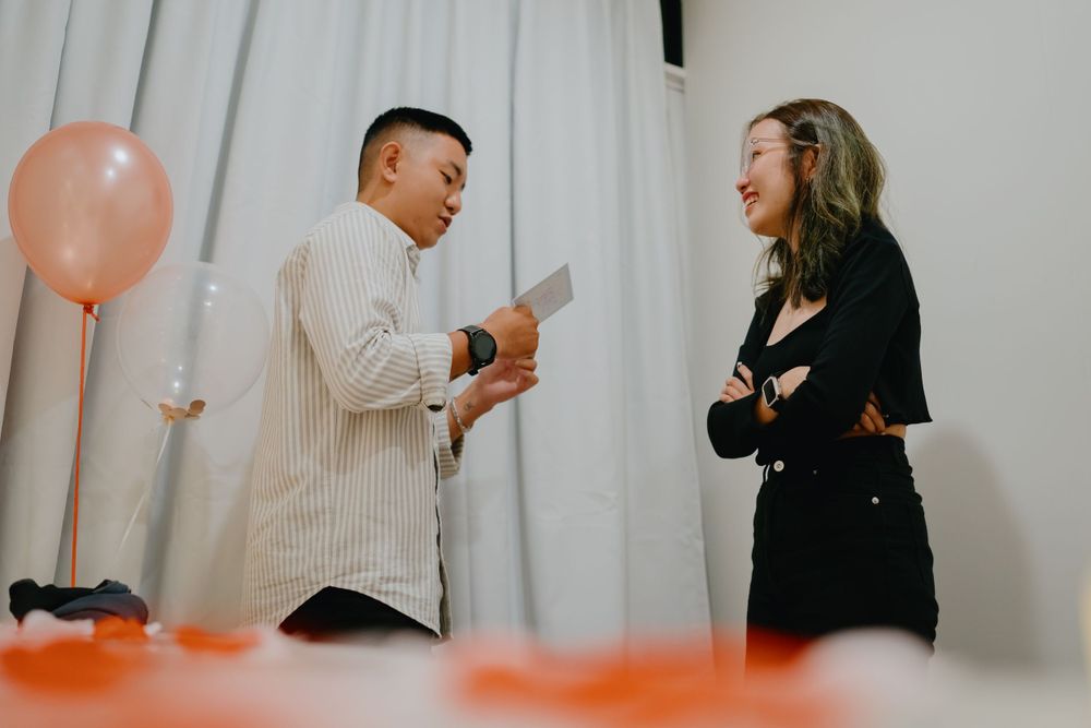 Two people laugh and interact at a casual event with orange balloons and white curtains in the background.