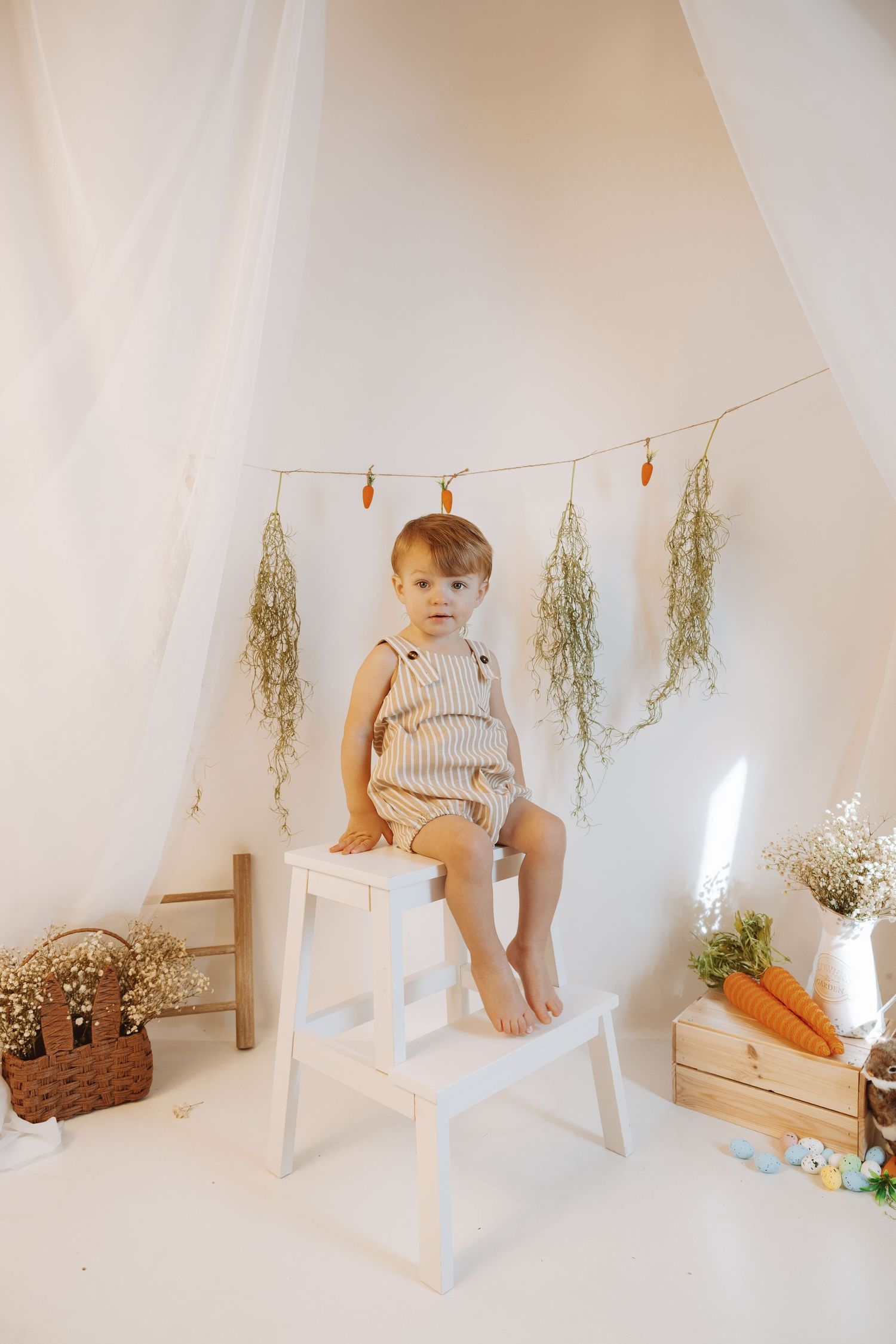 Small child in striped romper sits on white step stool with dried flowers and decor in minimalist studio setting.