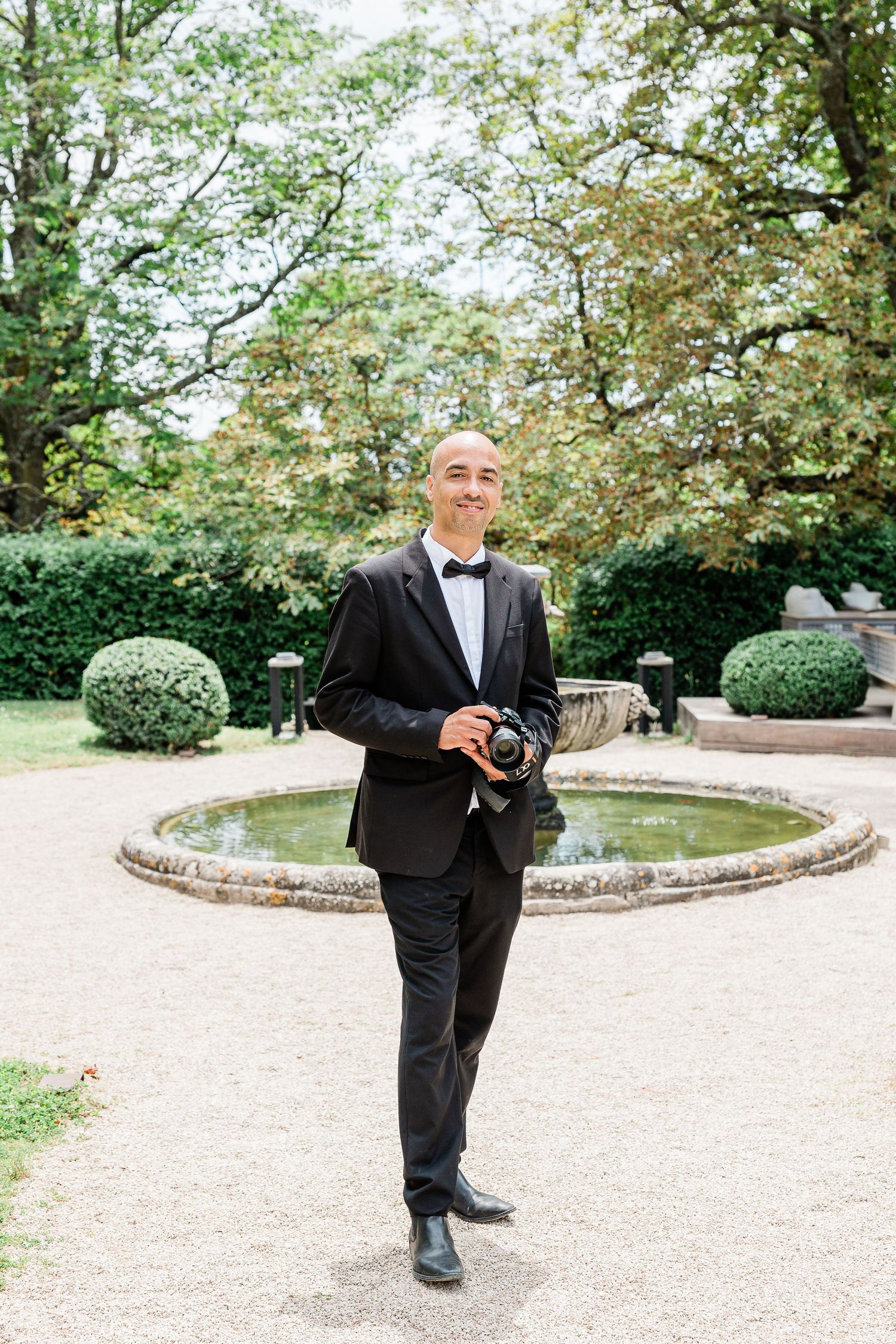 A professional photographer in formal black tuxedo stands at a garden courtyard with ornamental bushes and a water feature.