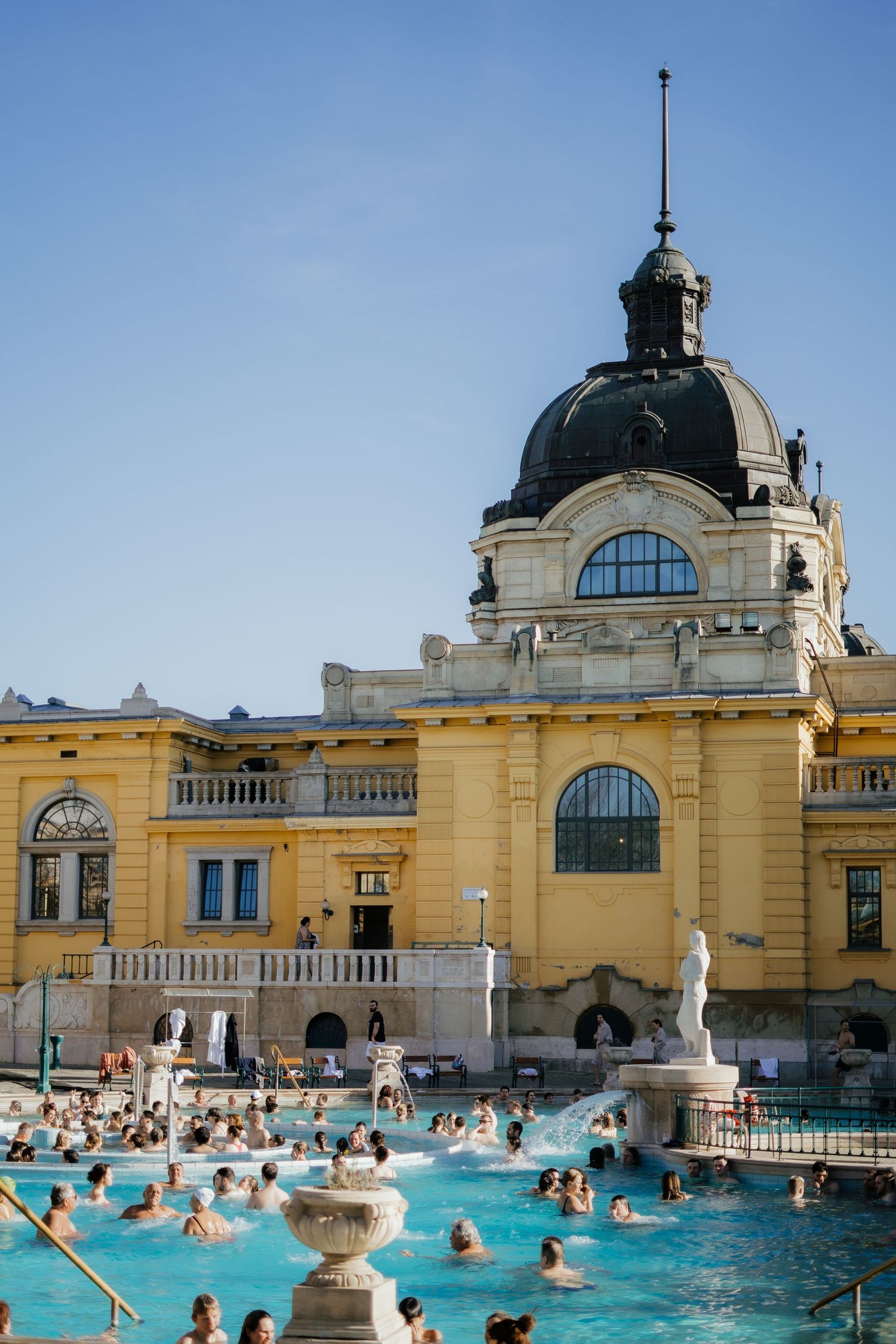People swim in the thermal baths at the historic Szechenyi Bath House with its yellow baroque architecture in Budapest.