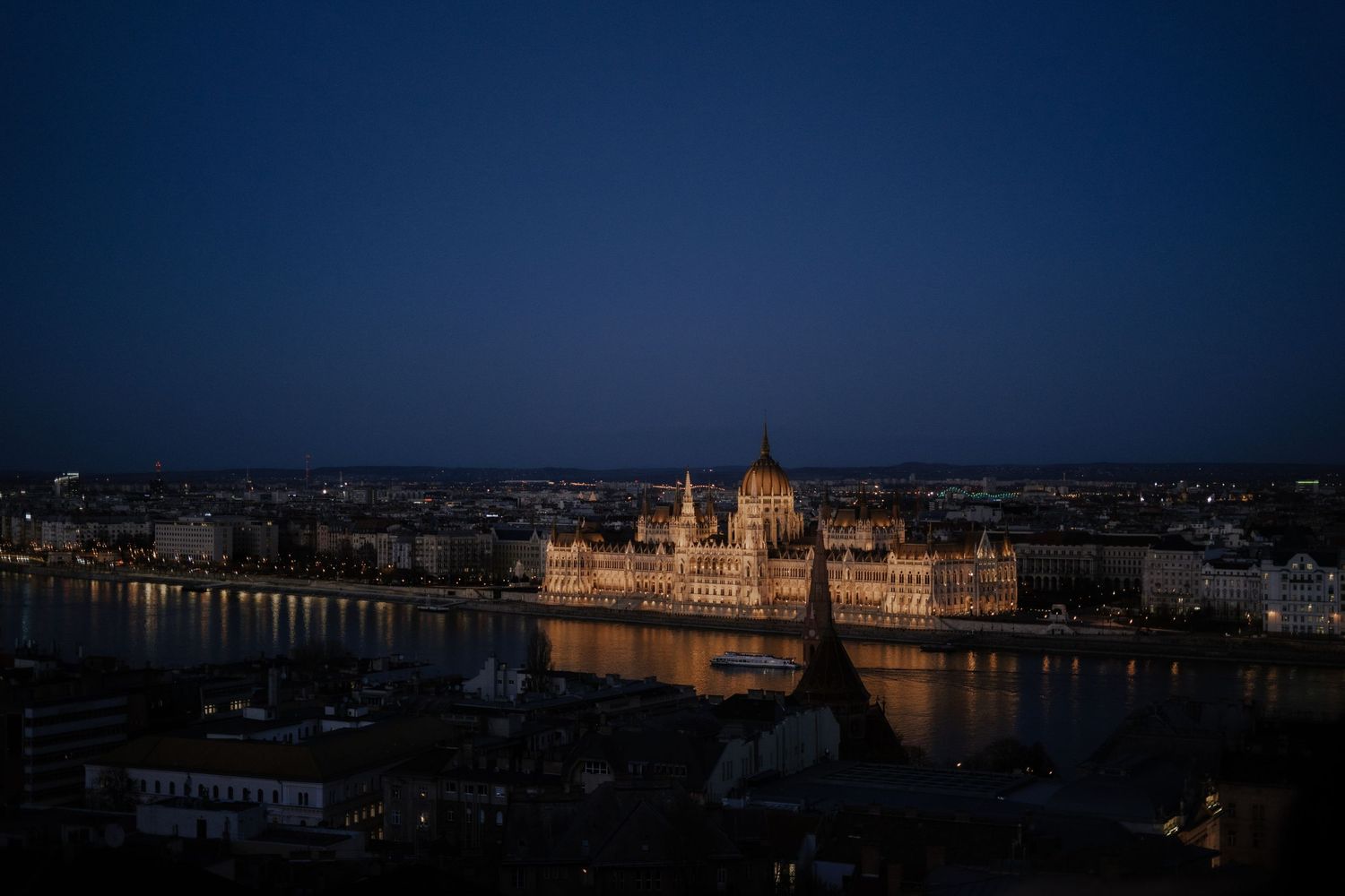 The illuminated Hungarian Parliament Building reflects in the Danube River at dusk in Budapest, creating a golden glow.