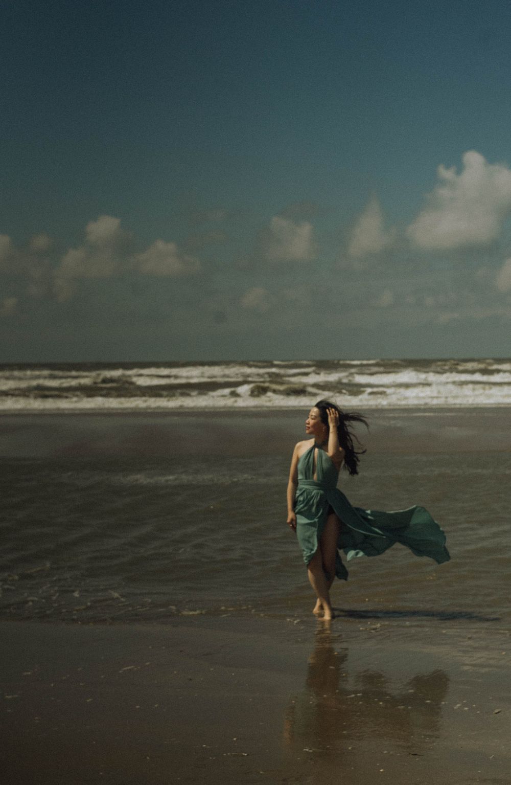 Person in flowing blue dress walking along a dramatic beach shoreline.