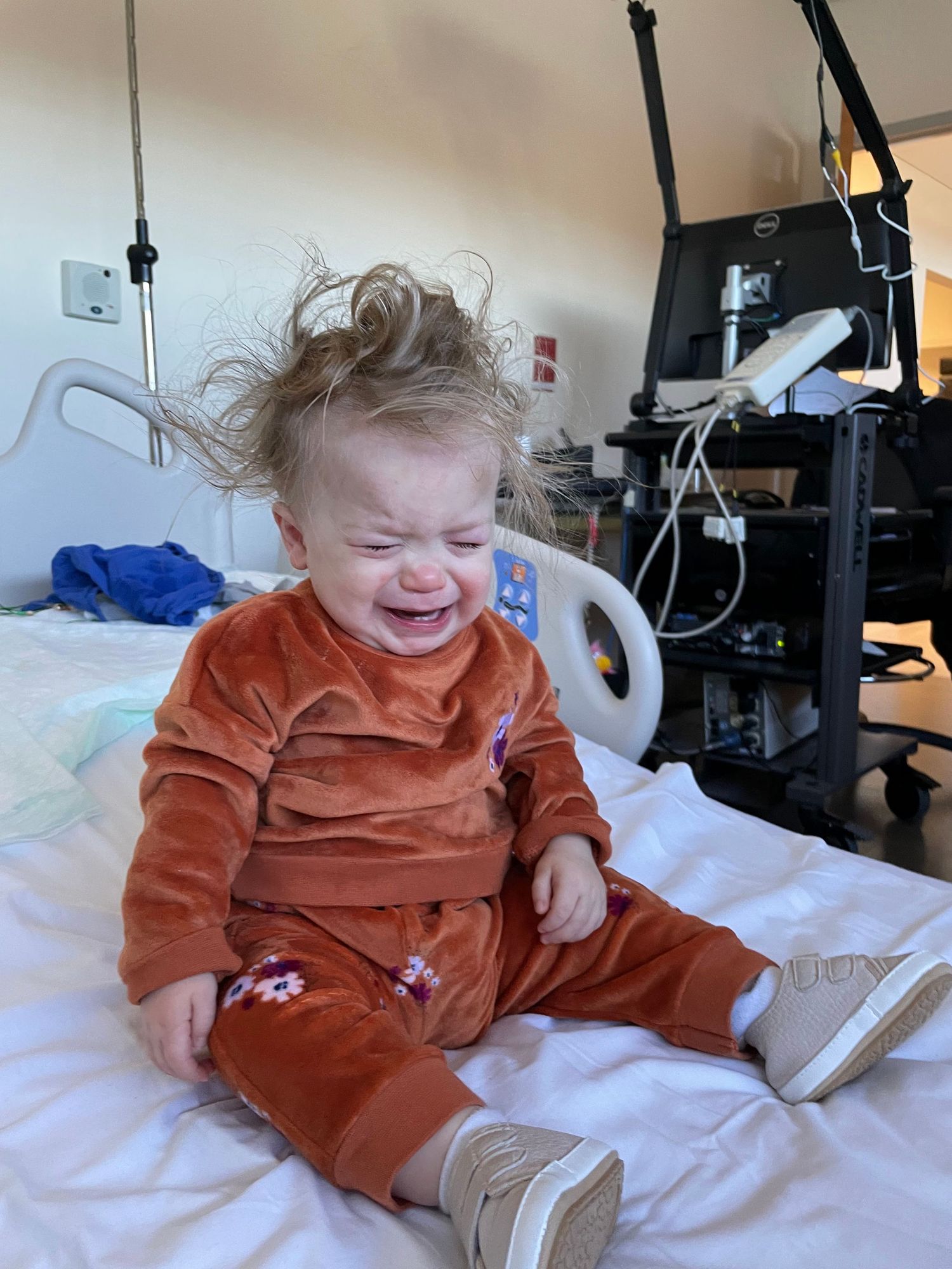 Baby in rust-colored velvet outfit sits on hospital bed with messy hair and expressive emotion.