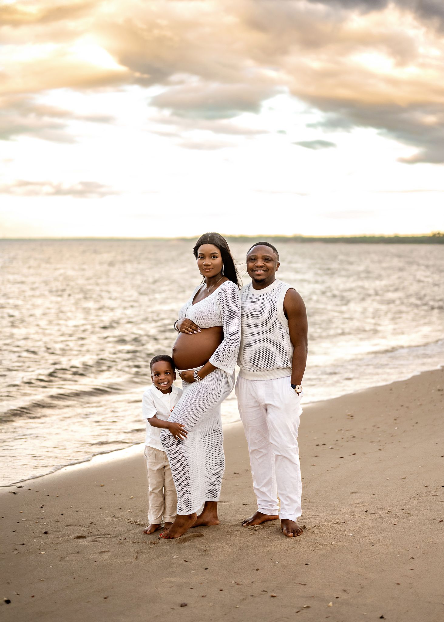 A family wearing white outfits poses together on a beach at sunset with golden clouds in the sky.