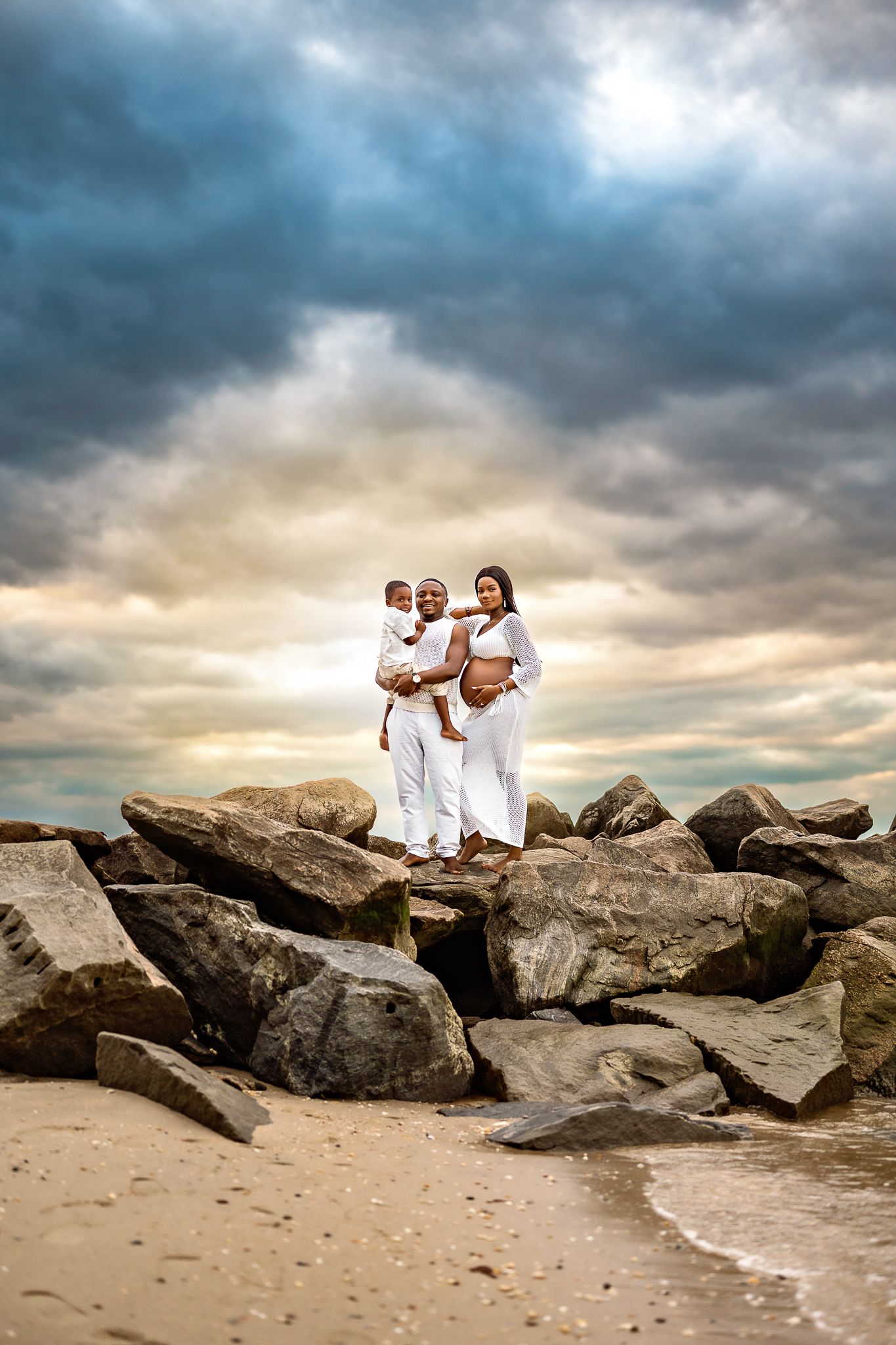 Family dressed in white poses together on rocky beach during dramatic sunset with moody clouds overhead.