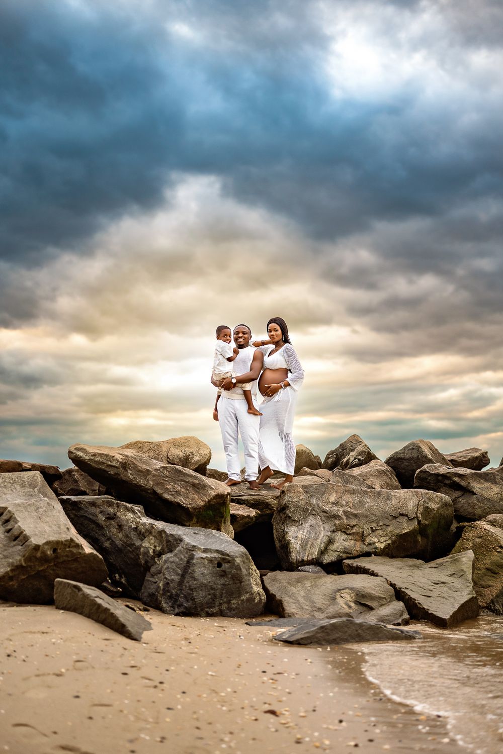 Family dressed in white poses together on rocky beach during dramatic sunset with moody clouds overhead.