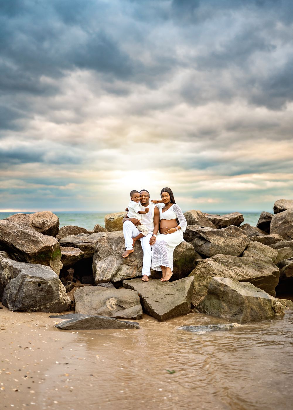 A family poses together on large rocks at a beach during sunset with dramatic cloudy skies overhead.