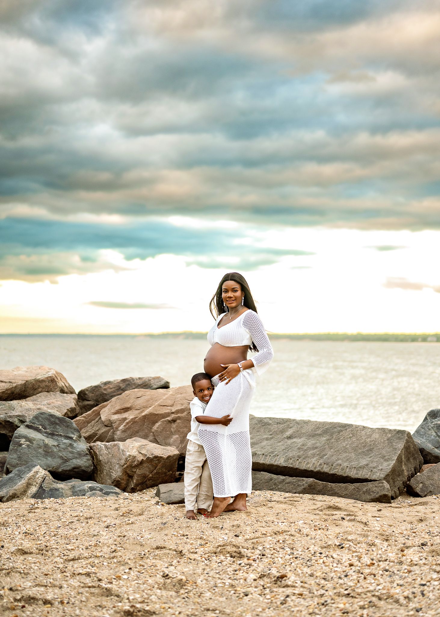 Parent and child in matching white outfits embrace on beach with dramatic cloudy sky and ocean backdrop.