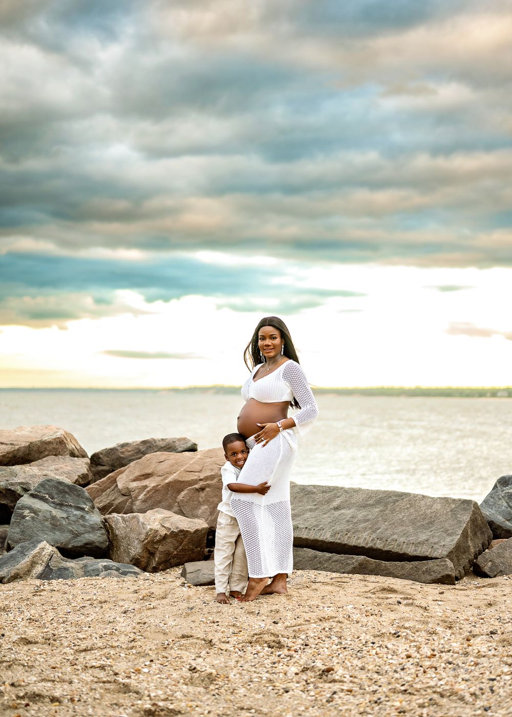 Parent and child in matching white outfits embrace on beach with dramatic cloudy sky and ocean backdrop.