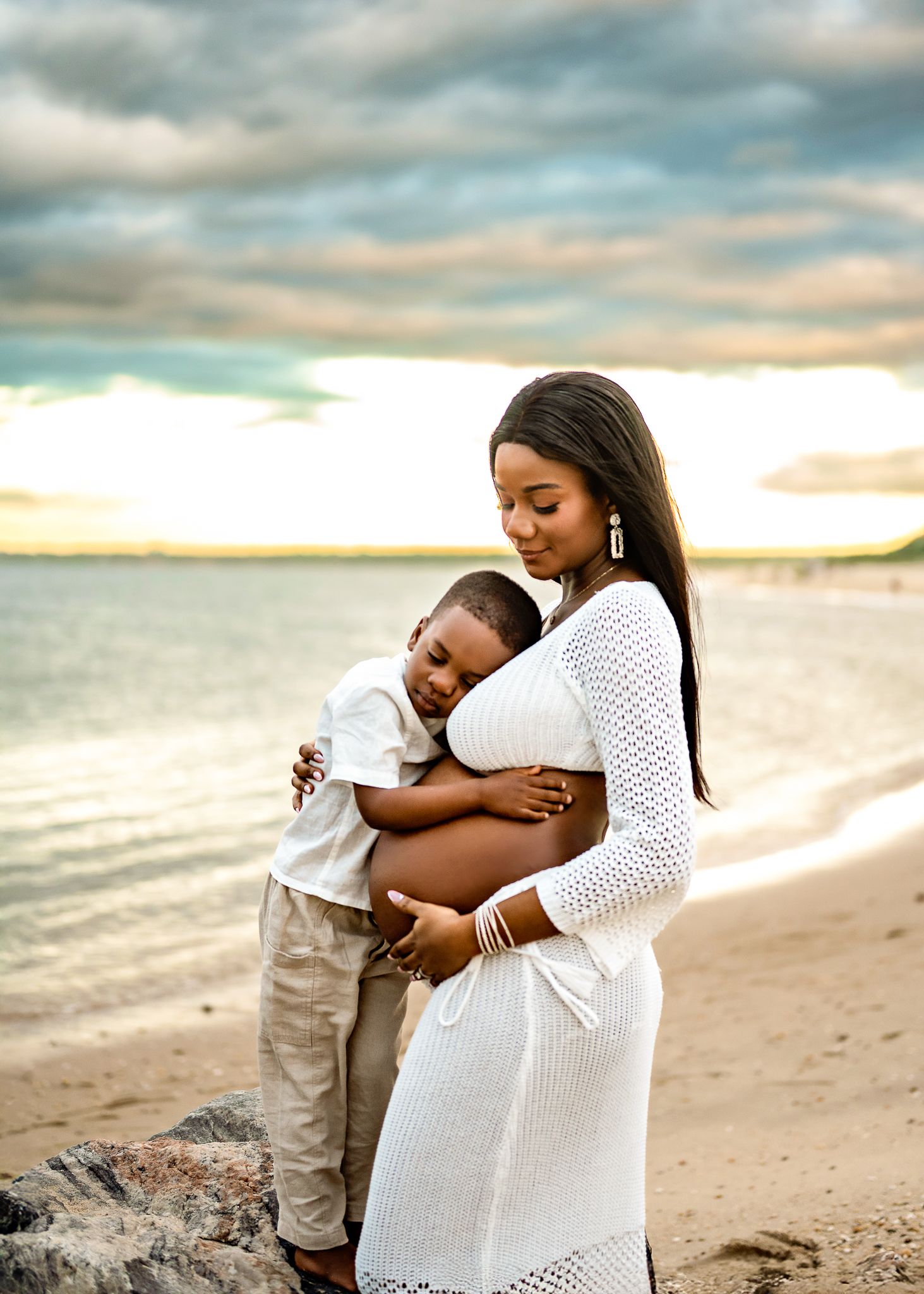 A mother and child share tender moments on a sandy beach at sunset, wearing matching white outfits.