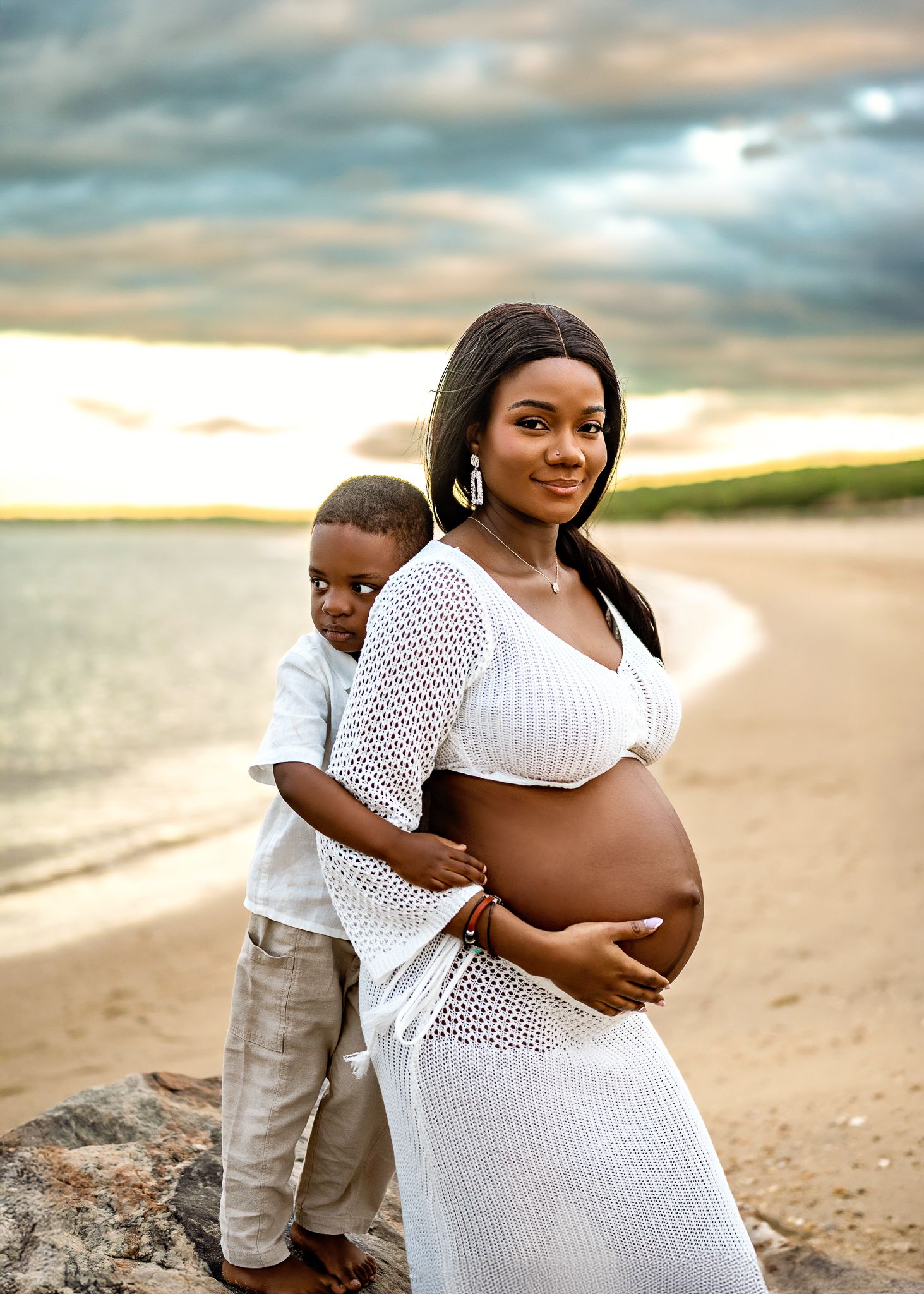 A serene beach maternity photo shows a glowing expectant mother in white attire with a companion embracing on the shore at sunset.