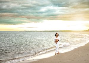 A person in white clothing walks along a serene beach at sunset with dramatic cloudy skies above the ocean.