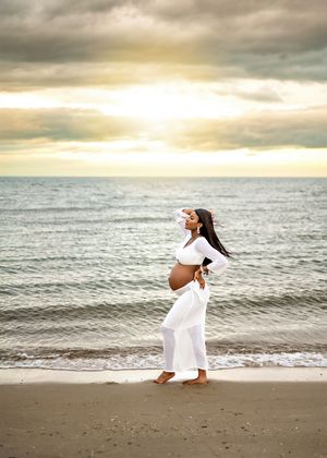 A figure in a white dress walks gracefully along a serene beach at sunset with dramatic cloudy skies above.