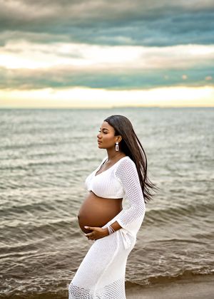 Expectant mother in flowing white dress poses serenely by the ocean during sunset with dramatic cloudy sky backdrop.