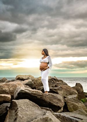 Pregnant woman in white outfit stands gracefully on coastal rocks at sunset with dramatic cloudy sky overhead.