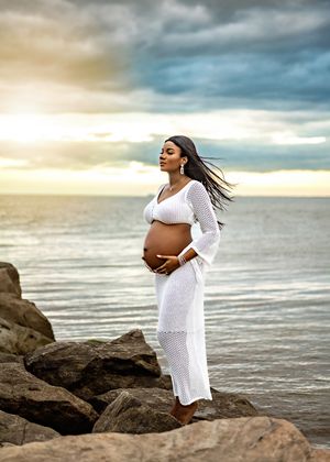 A pregnant woman in a flowing white dress stands peacefully on coastal rocks at sunset while her hair blows in the breeze.