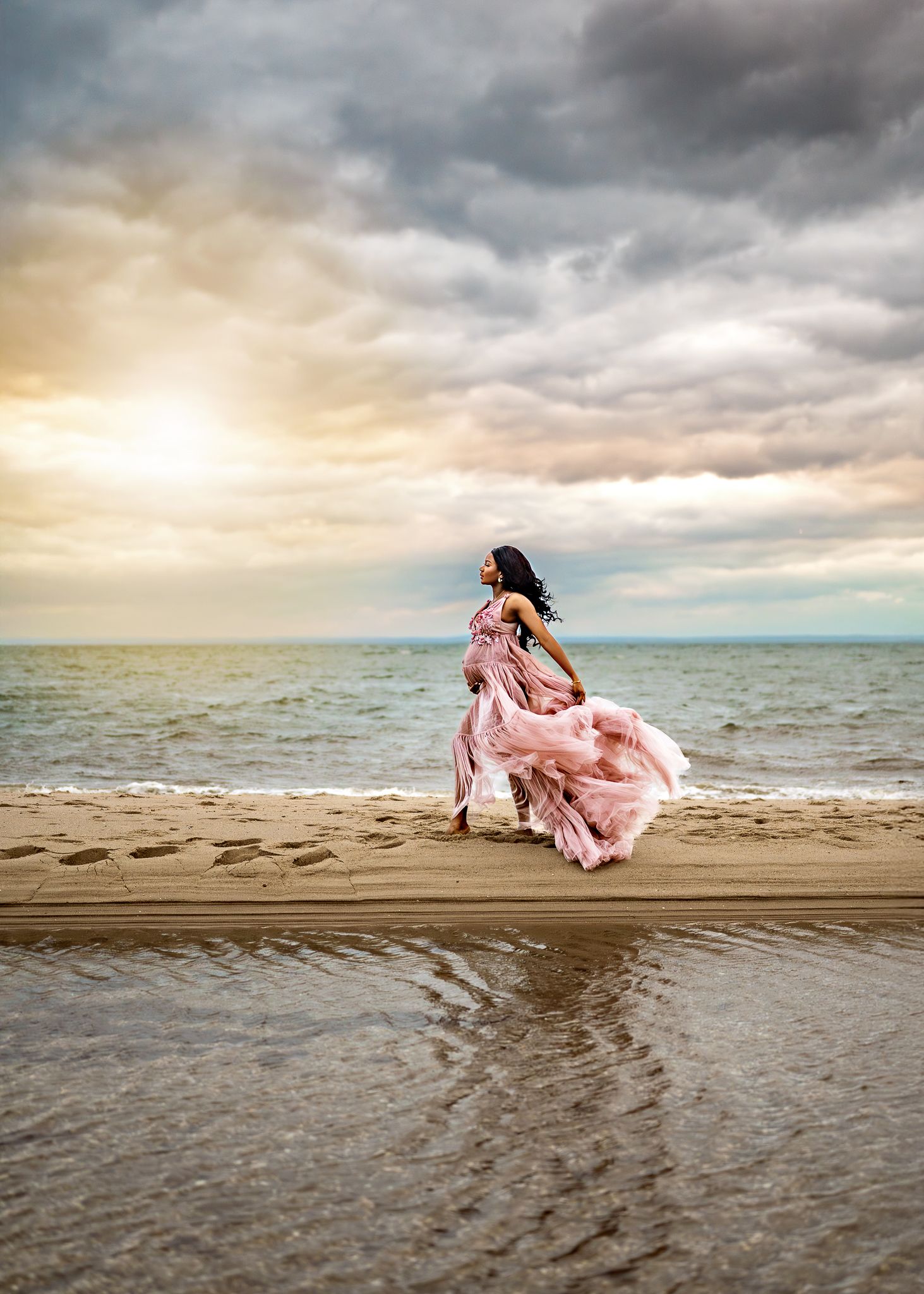 A figure in a flowing pink dress twirls gracefully on a beach at sunset with dramatic stormy clouds overhead.