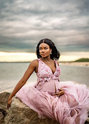 A person in a flowy pink gown sits gracefully on coastal rocks at sunset beneath dramatic cloudy skies.