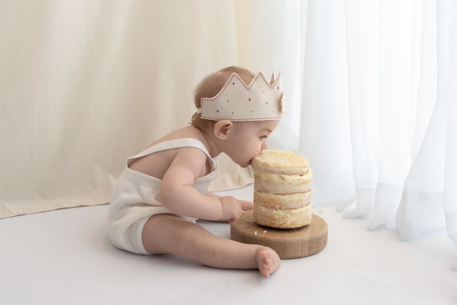Baby wearing a crown sits near a naked cake on a wooden stand against a white backdrop.