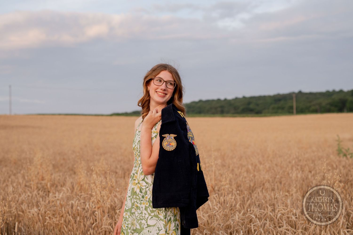 Person in FFA jacket and floral dress poses in golden wheat field at sunset.