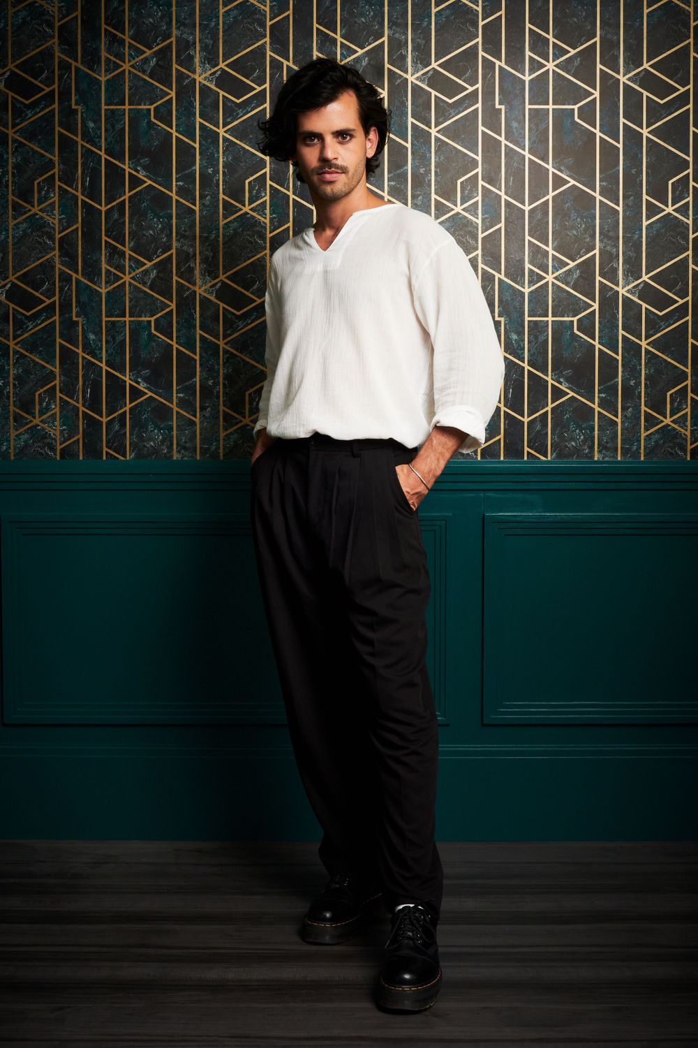 A person in a casual white sweater and black pants poses against a dark green wall with gold geometric patterns.