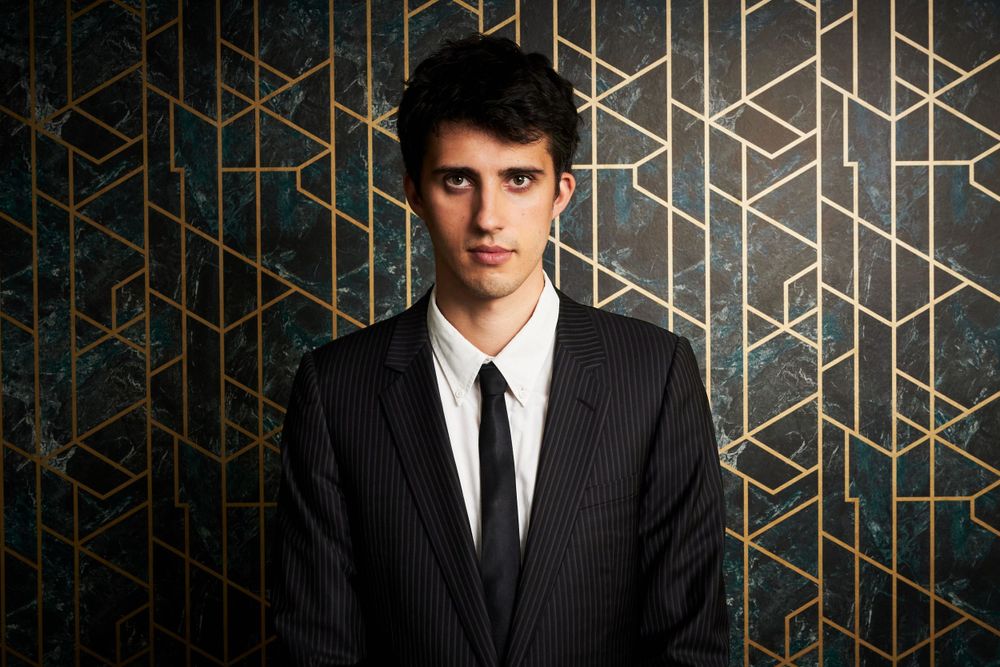 Professional portrait of a person in a pinstriped suit and black tie against a modern geometric gold patterned background.