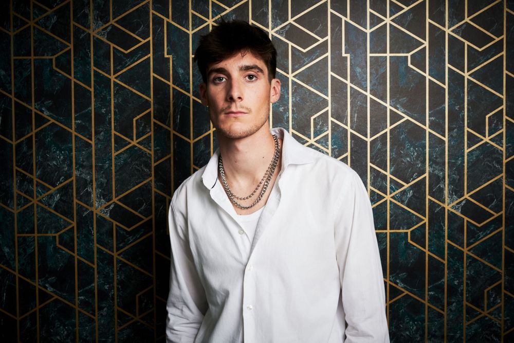 A person in a white button-down shirt and gold necklace poses against a geometric patterned wall.