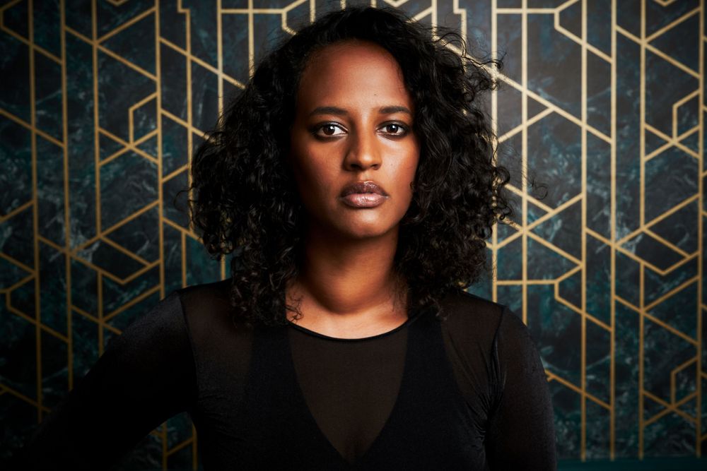 Portrait of a person with natural curly hair wearing black against an art deco patterned green and gold wall.