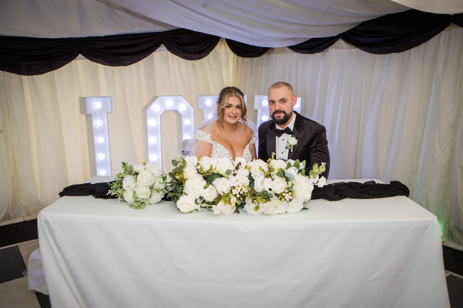 Wedding reception table decorated with white floral arrangements and illuminated LOVE sign.