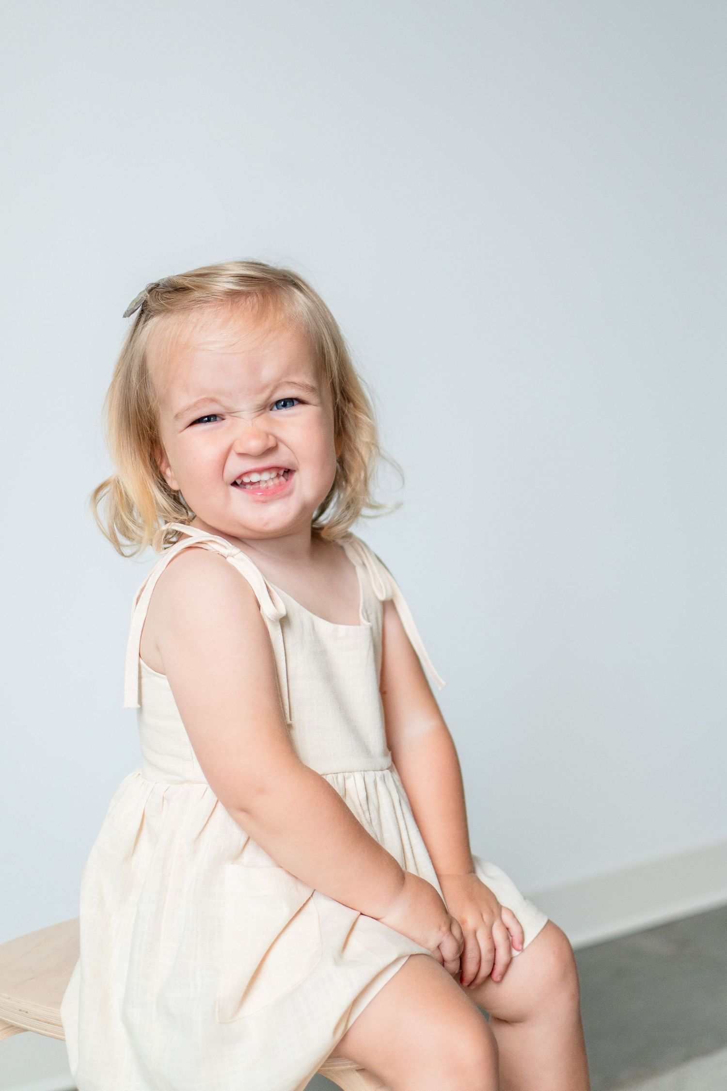 A small child in a cream-colored dress sits and smiles joyfully against a light gray background.