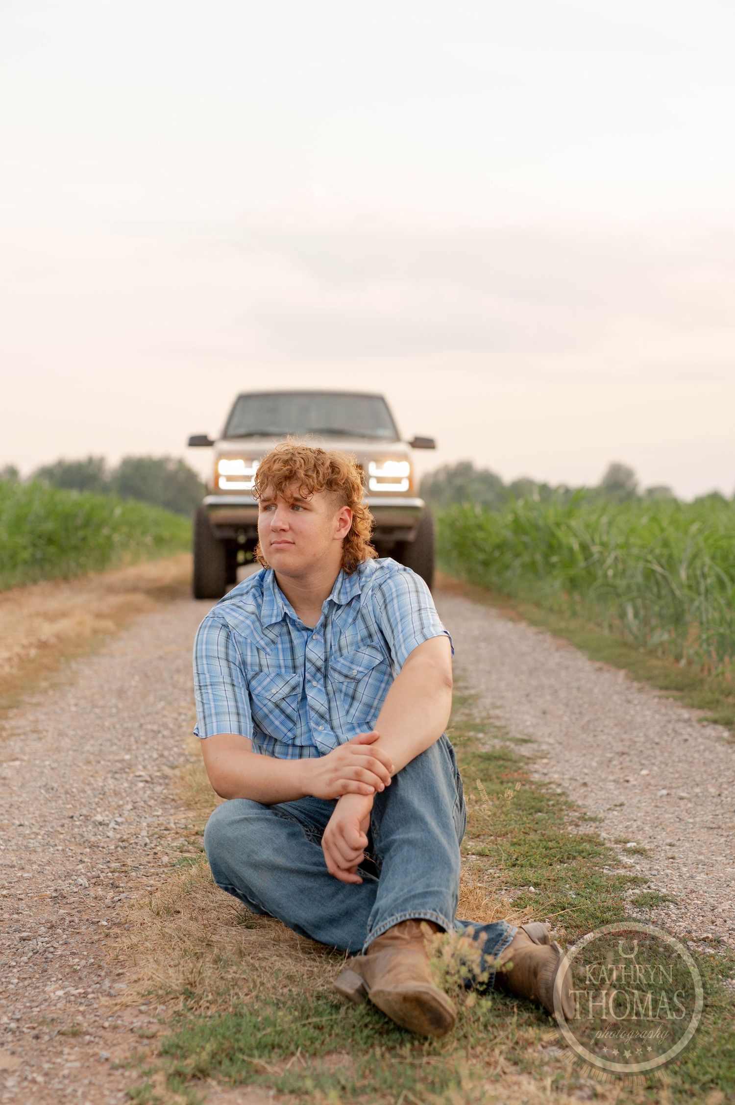 A person in a plaid shirt sits on a dirt road with a truck visible in the background.