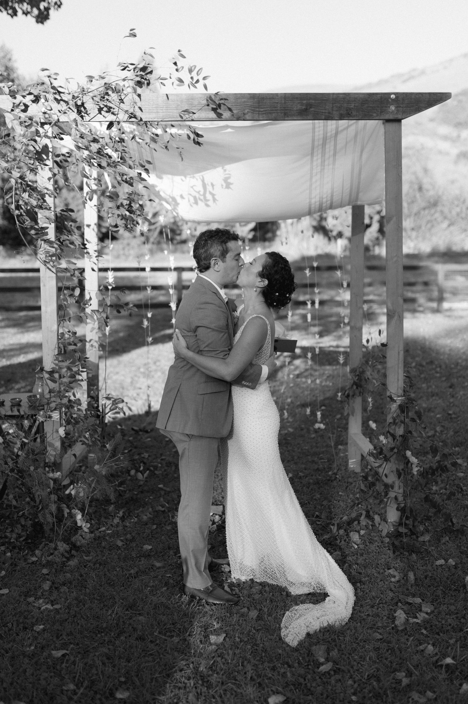 A romantic black and white wedding moment under a wooden arbor surrounded by trees.