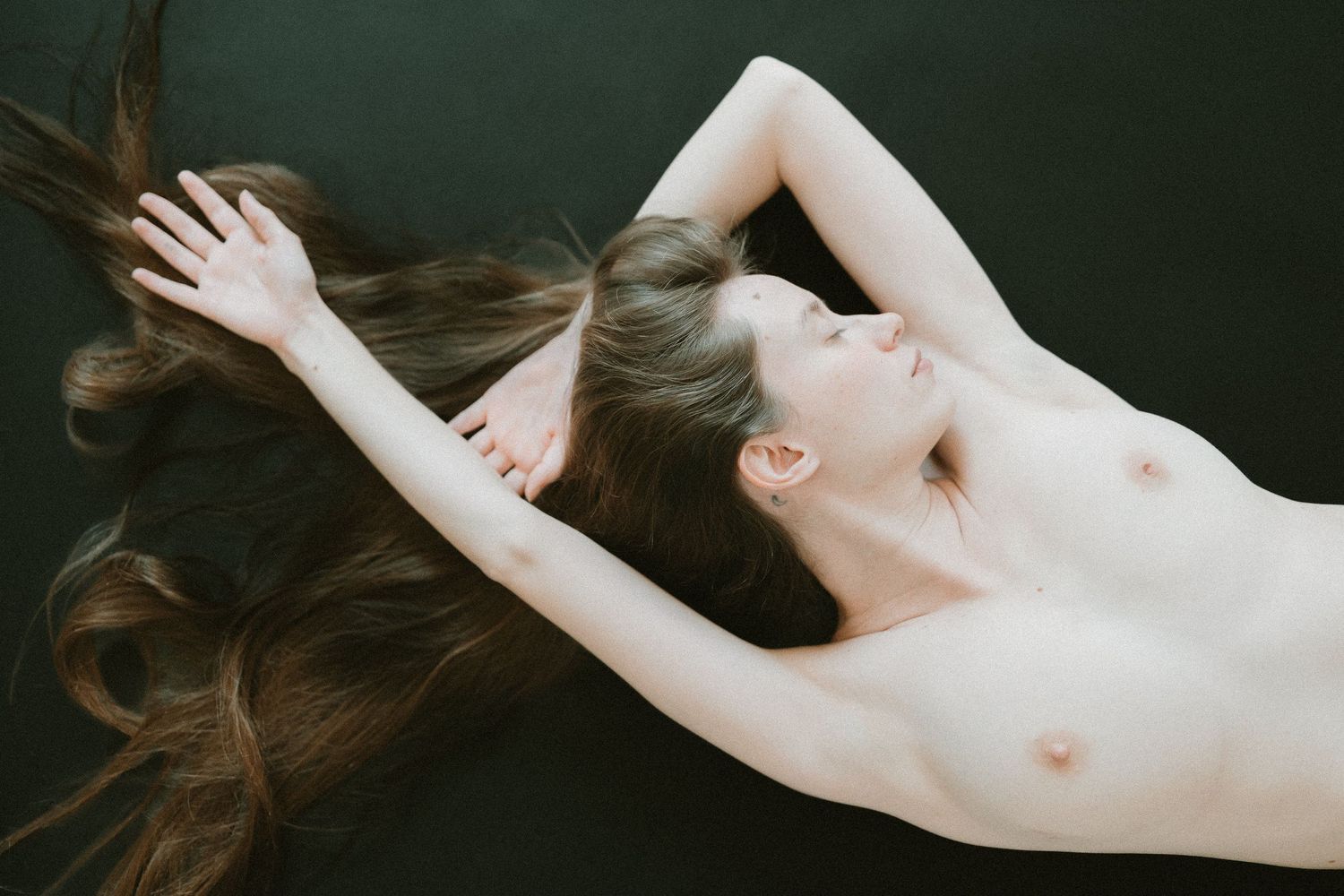 Artistic topless figure with long flowing brown hair lying against dark background in contemplative pose.