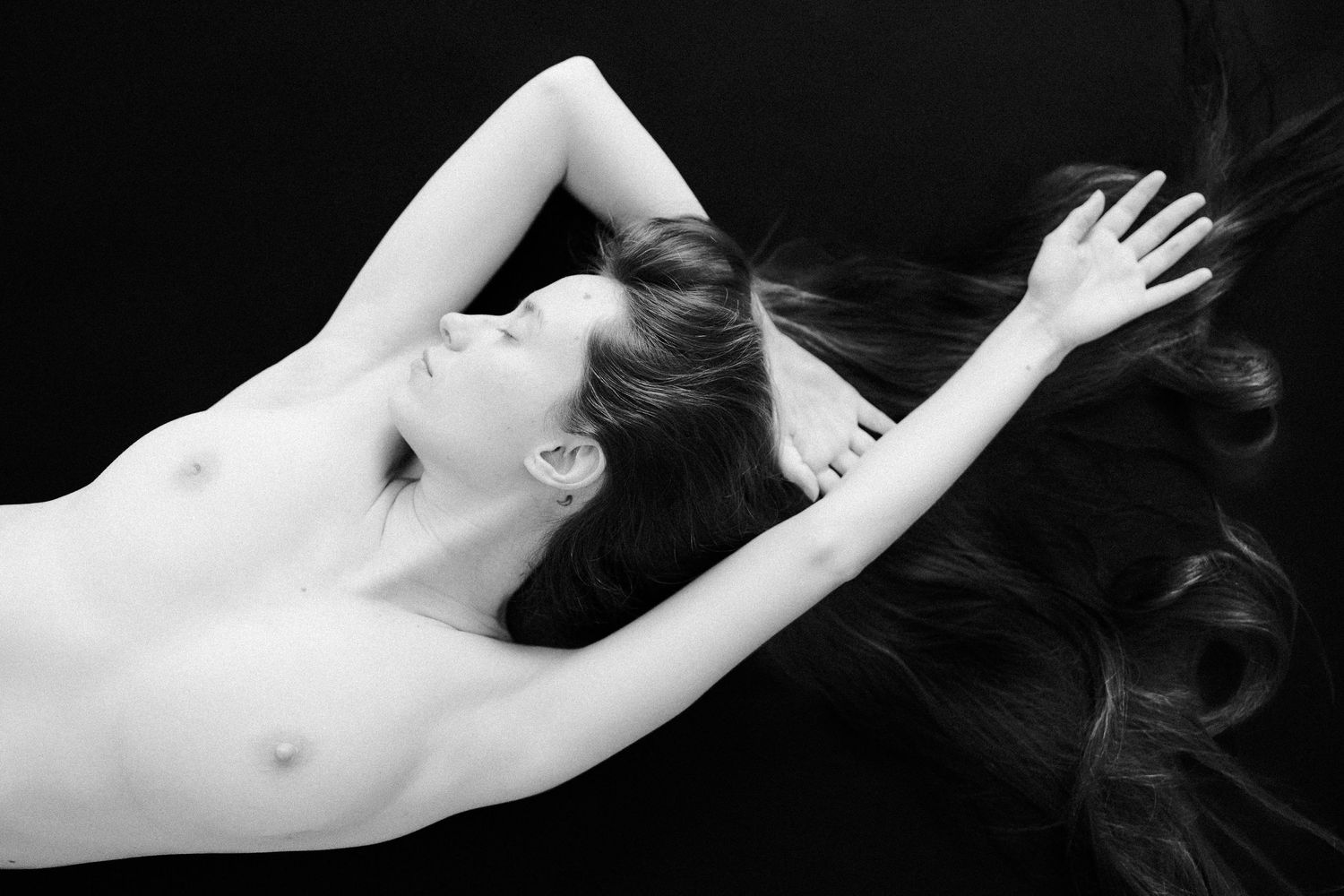 A black and white art nude photograph showing a reclining figure with dramatic lighting and flowing dark hair.