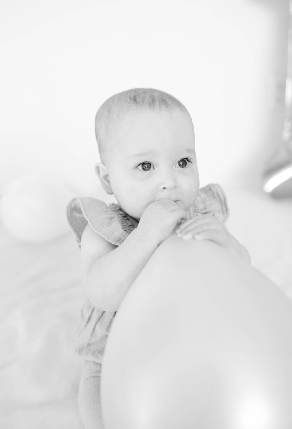 Black and white close-up photograph of a baby in a white dress looking upward while chewing their finger.