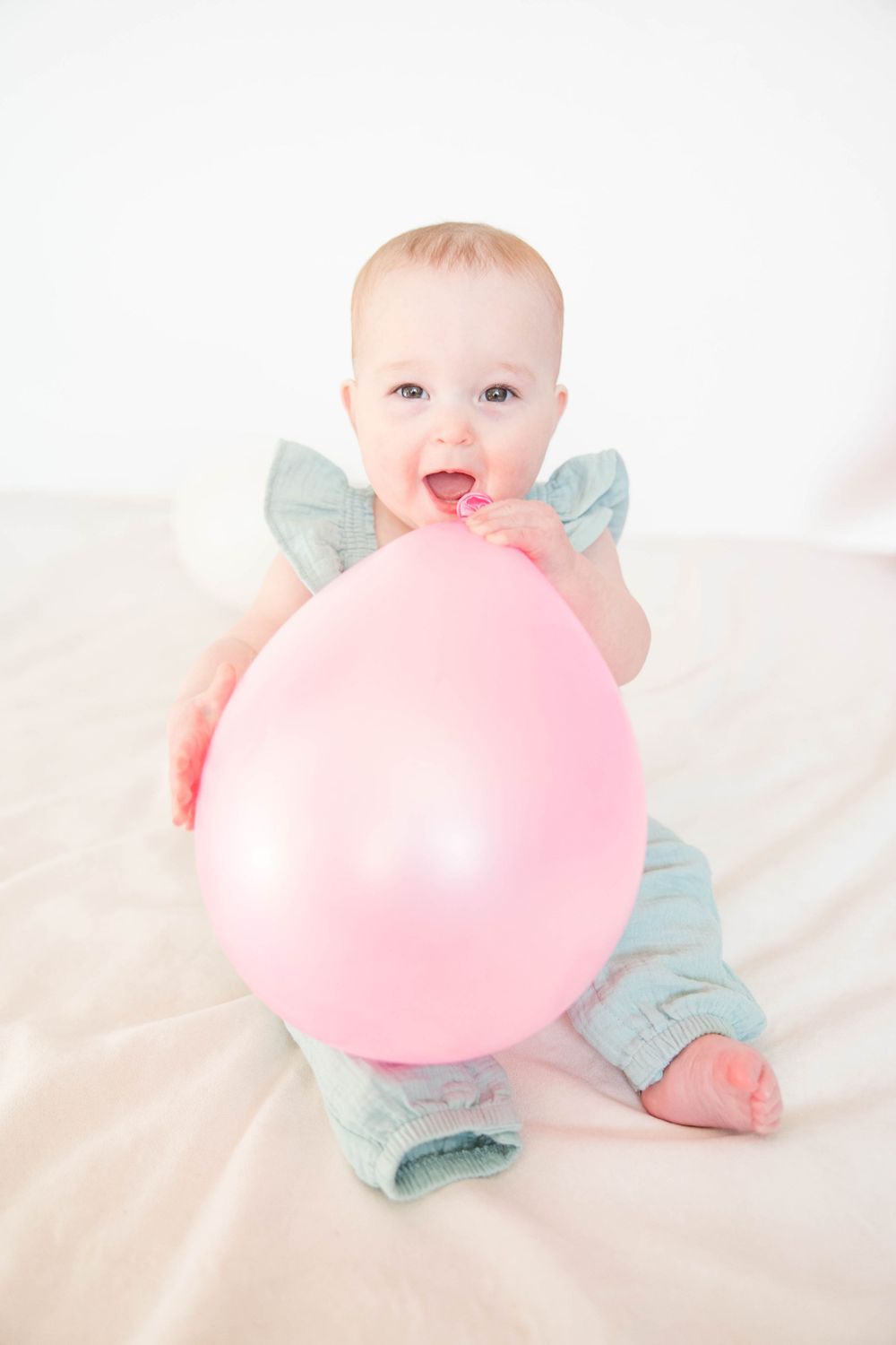Baby in mint green outfit plays with large pink balloon while sitting on white background.