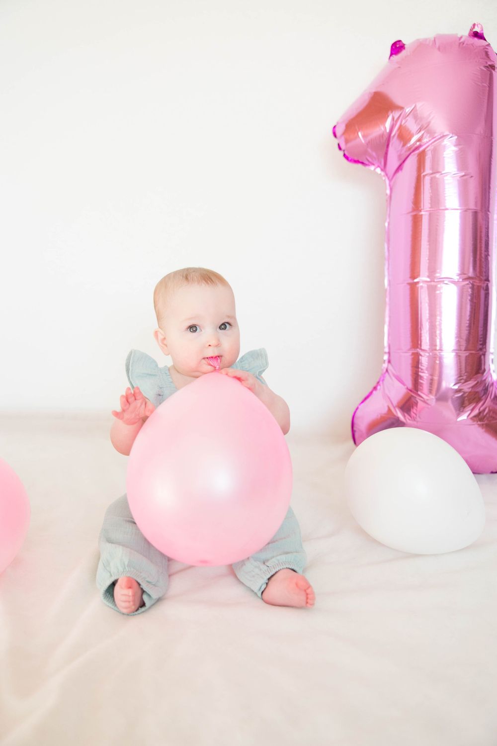 A baby in blue outfit sits with pink balloons and a large pink number one balloon for a first birthday photoshoot.