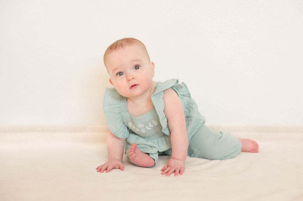 Baby wearing mint green romper crawls on light-colored studio backdrop during portrait session.