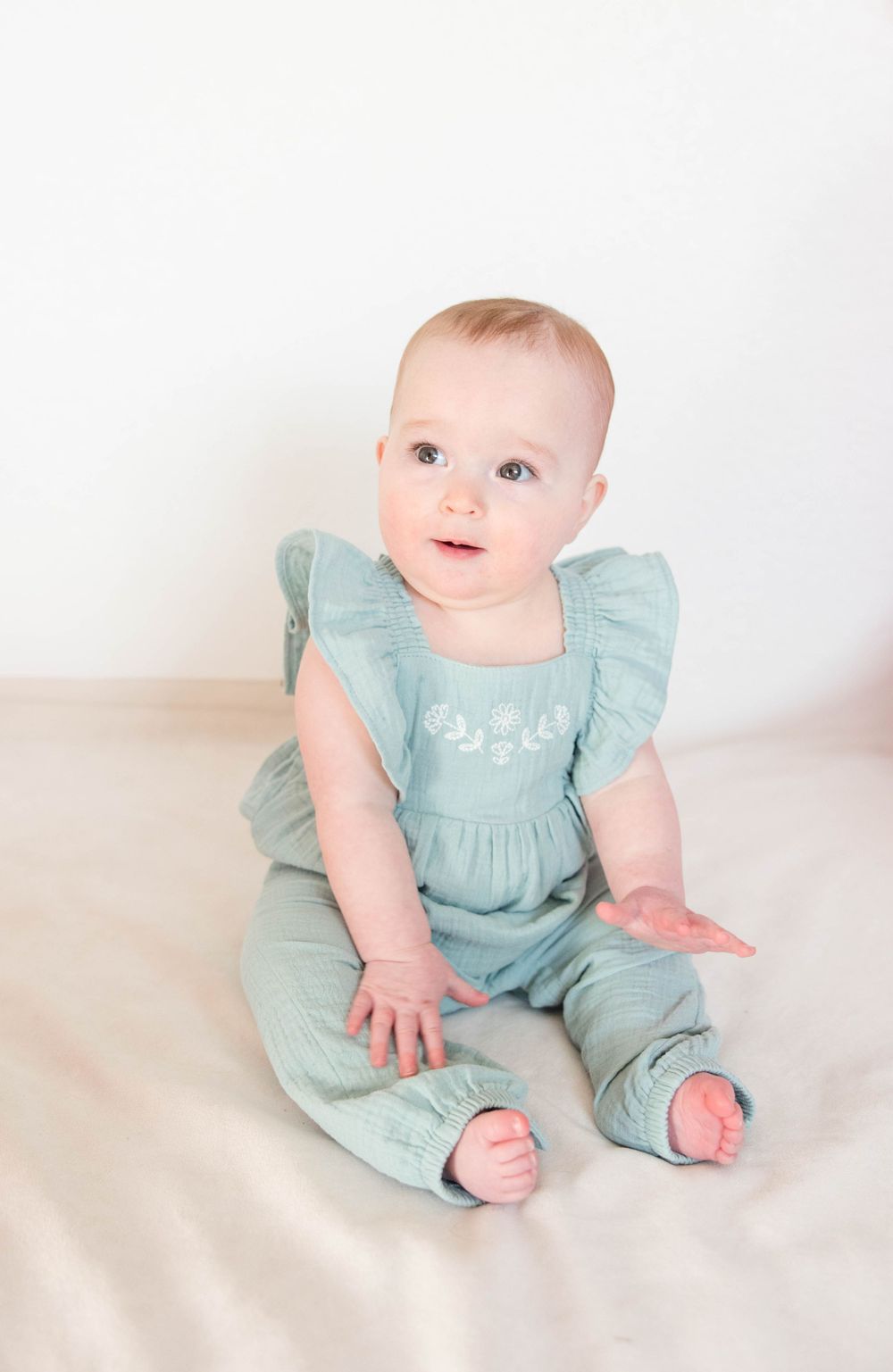 Baby wearing mint green ruffle romper sits on cream colored backdrop.