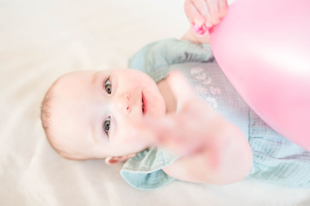 Baby in light blue outfit lying down with soft pink balloon against light background.
