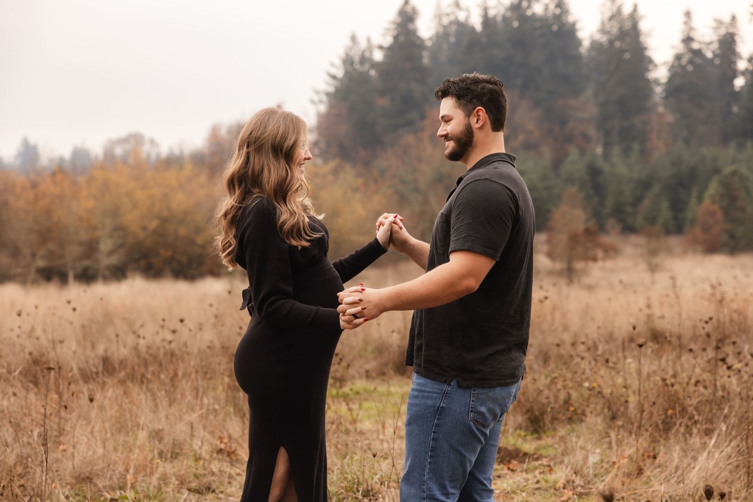 A couple stands facing each other in a golden dry grass field with evergreen trees in the background at sunset.