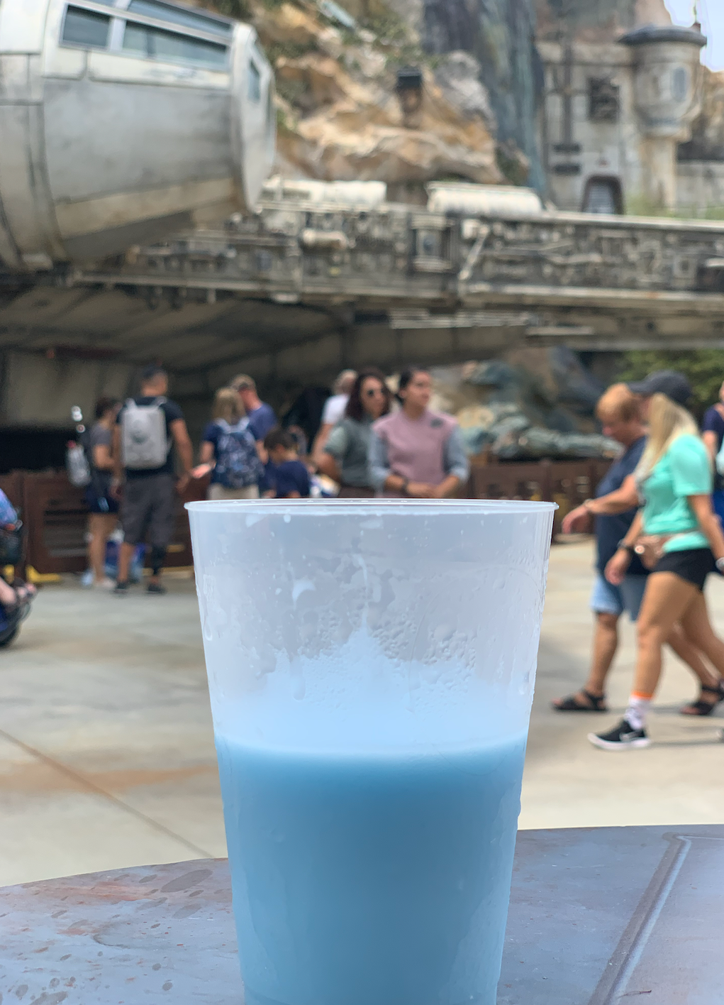A frosty blue beverage in a clear plastic cup with crowds visible at a theme park attraction.
