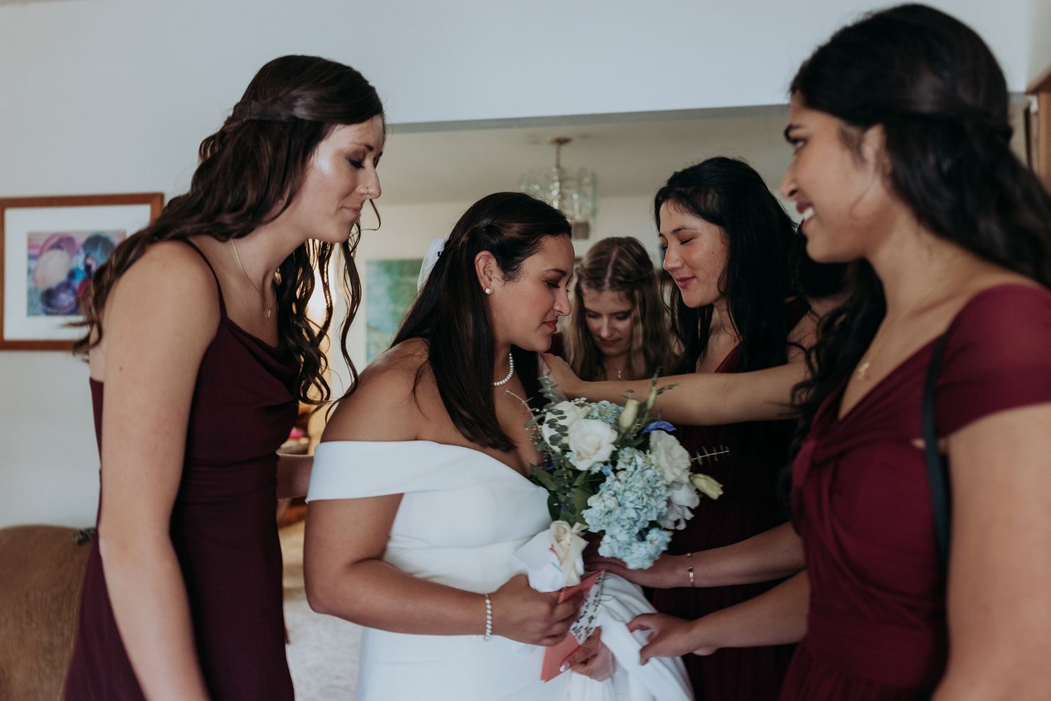 Bridesmaids in burgundy dresses help bride prepare for wedding ceremony in intimate getting ready moment.