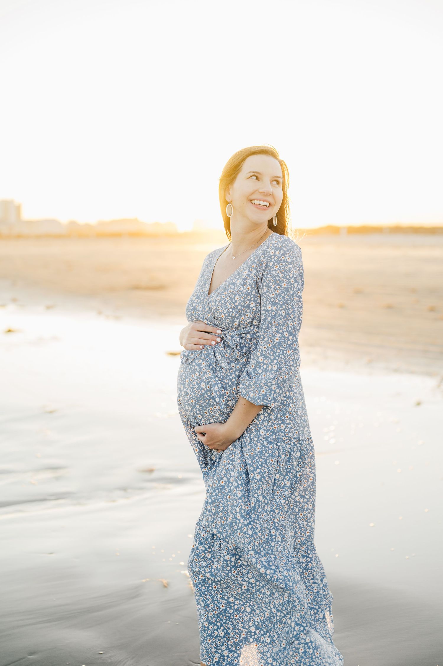 A woman in a blue floral dress poses on a sandy beach during golden hour sunset.