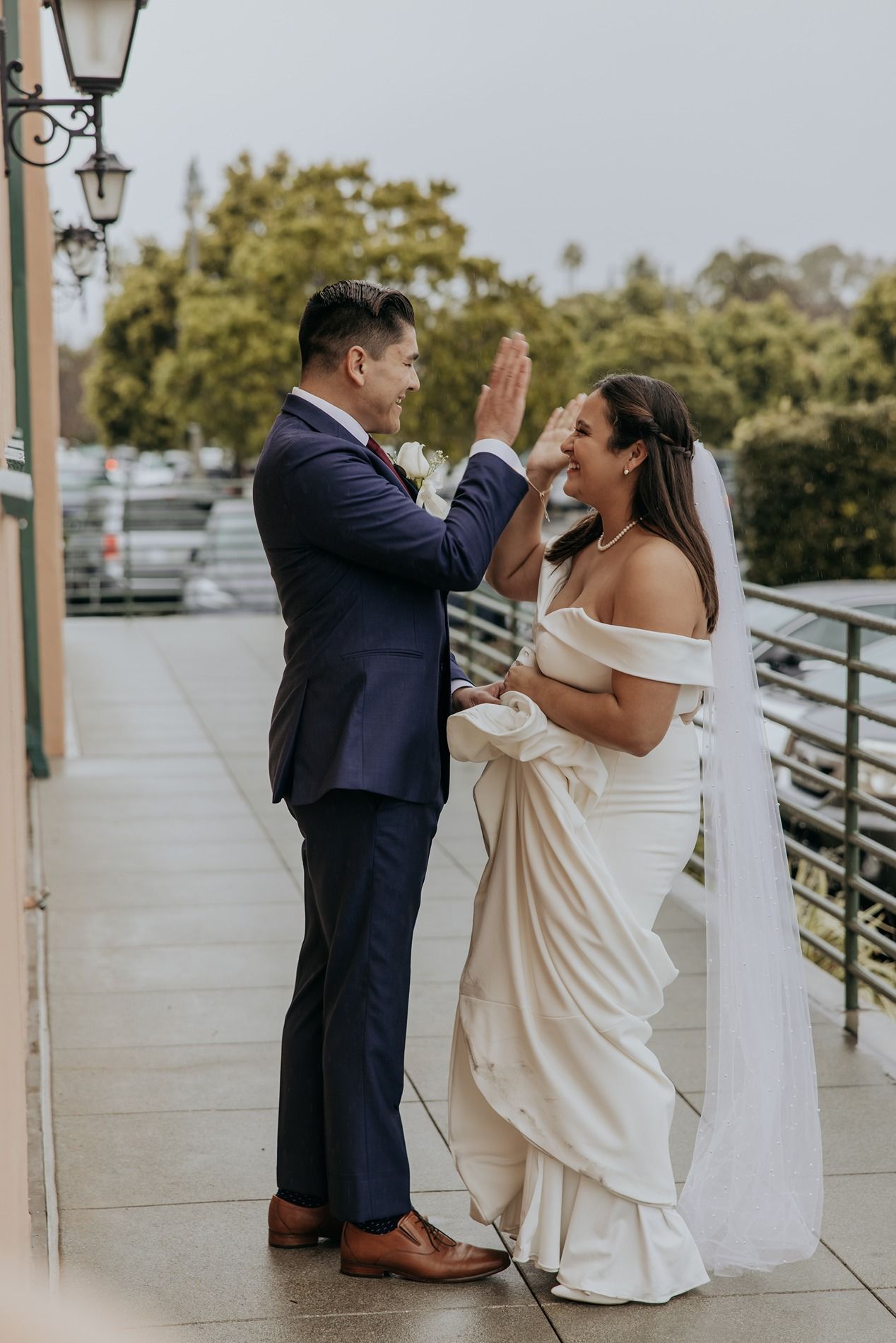 A couple in wedding attire shares a playful moment on a porch with lamp posts and railings at BRICK Liberty Station.