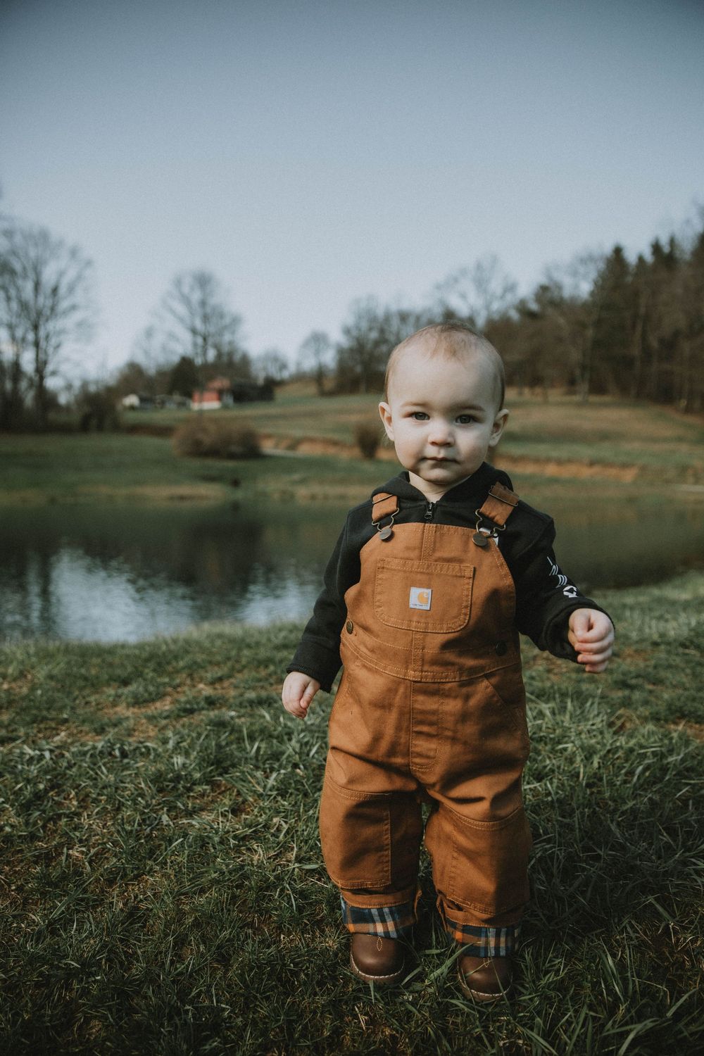 A small child wearing brown overalls stands in a grassy field with a pond and trees in the background on a cloudy day.