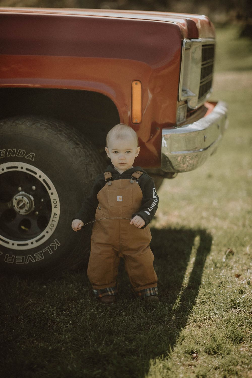 Small figure in brown overalls stands next to vintage red pickup truck with rugged off-road tires on grass.